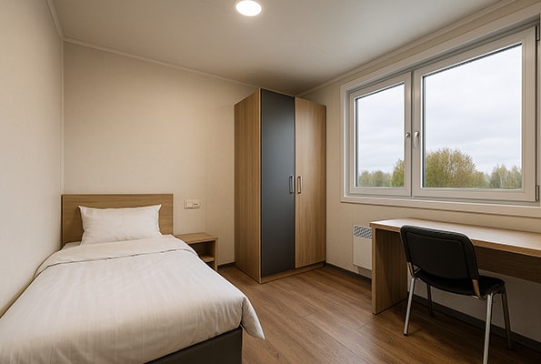 Jednolůžkový pokoj v kontejneru šířky ~2,4 m – postel, skříň a pracovní stůl.